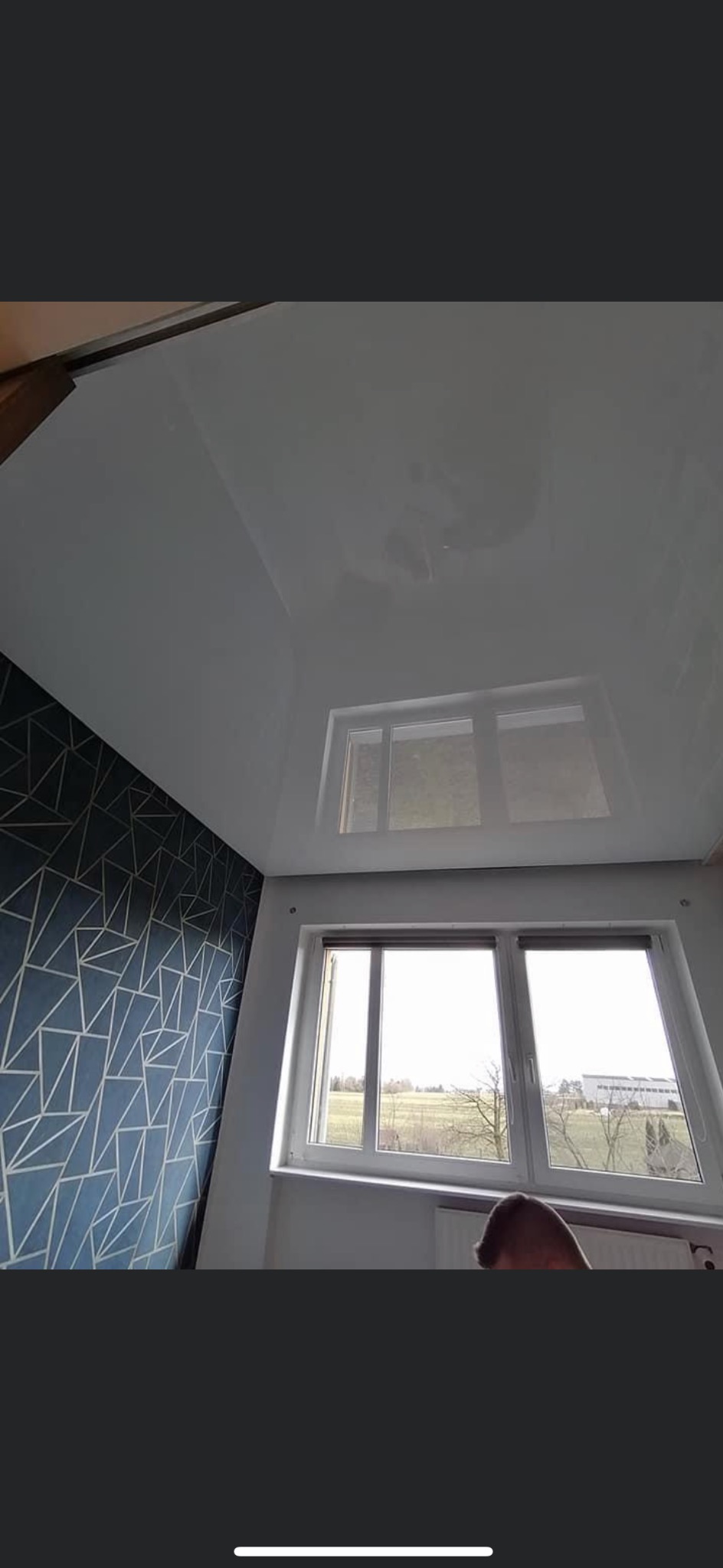 Biały, błyszczący sufit napinany z odbiciem okna, zestawiony z niebieską ścianą w geometryczne wzory, widok z dołu.