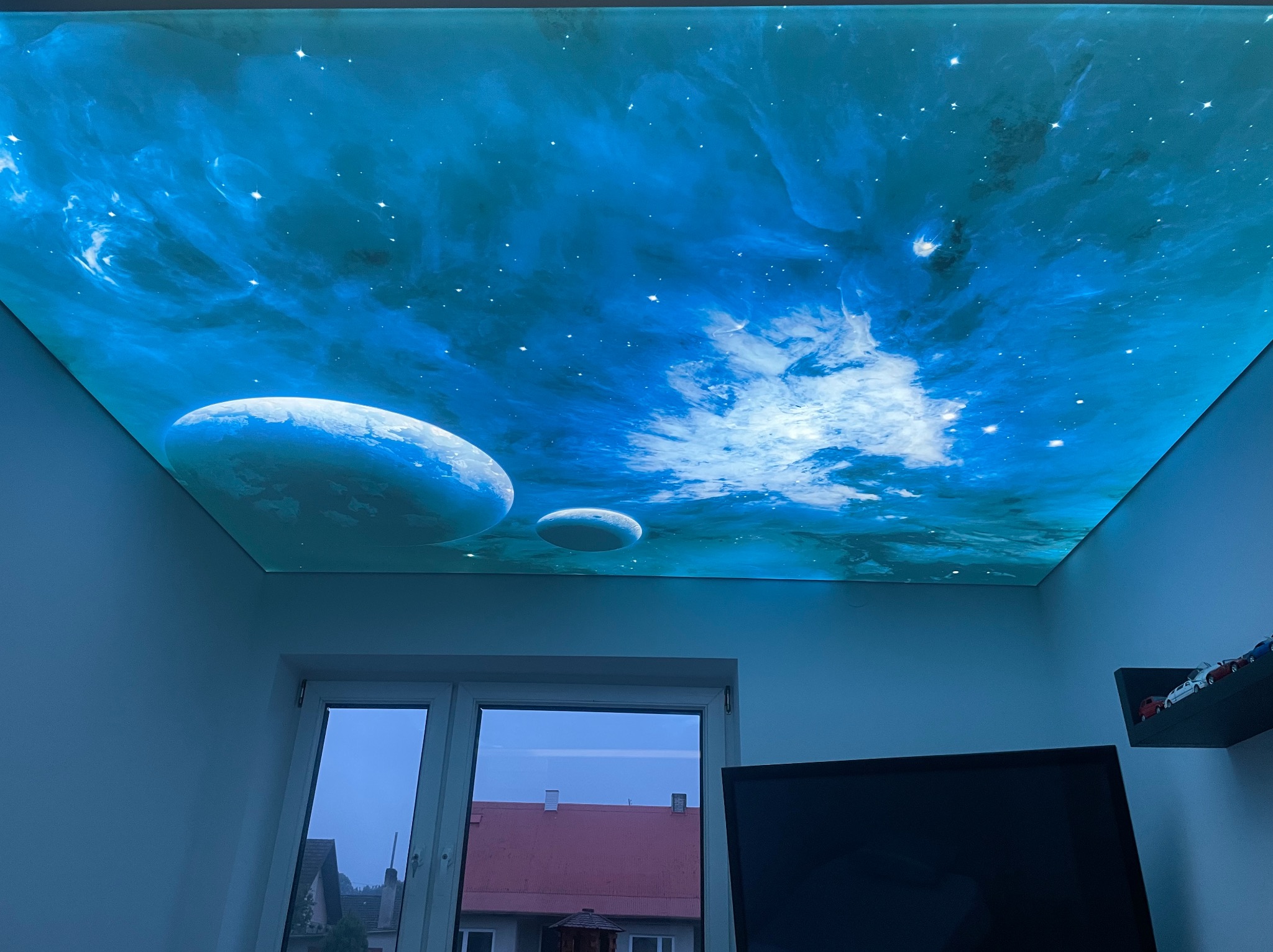 Podświetlany sufit napinany z motywem kosmosu: planety i mgławice w odcieniach błękitu, widoczne z perspektywy pokoju z oknem i telewizorem.