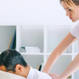 Kobieta w białej koszulce wykonuje masaż pleców mężczyźnie leżącemu na białym stole w jasnym gabinecie.