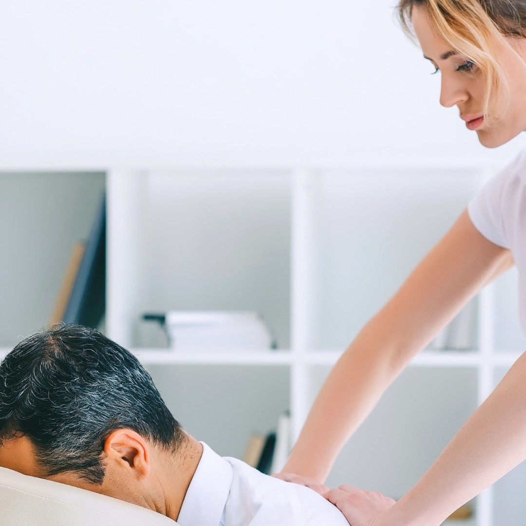 Kobieta w białej koszulce wykonuje masaż pleców mężczyźnie leżącemu na białym stole w jasnym gabinecie.