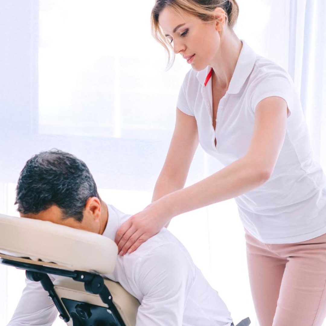 Kobieta w białej koszuli wykonuje relaksacyjny ucisk karku na mężczyźnie siedzącym na specjalistycznym krześle do sesji w biurze, jasne tło.