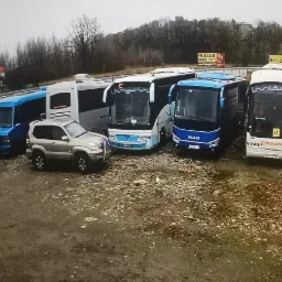Flota pojazdów transportowych, w tym autobusy MAN i mniejsze busy, zaparkowane na żwirowym placu z widoczną reklamą billboardową w tle.