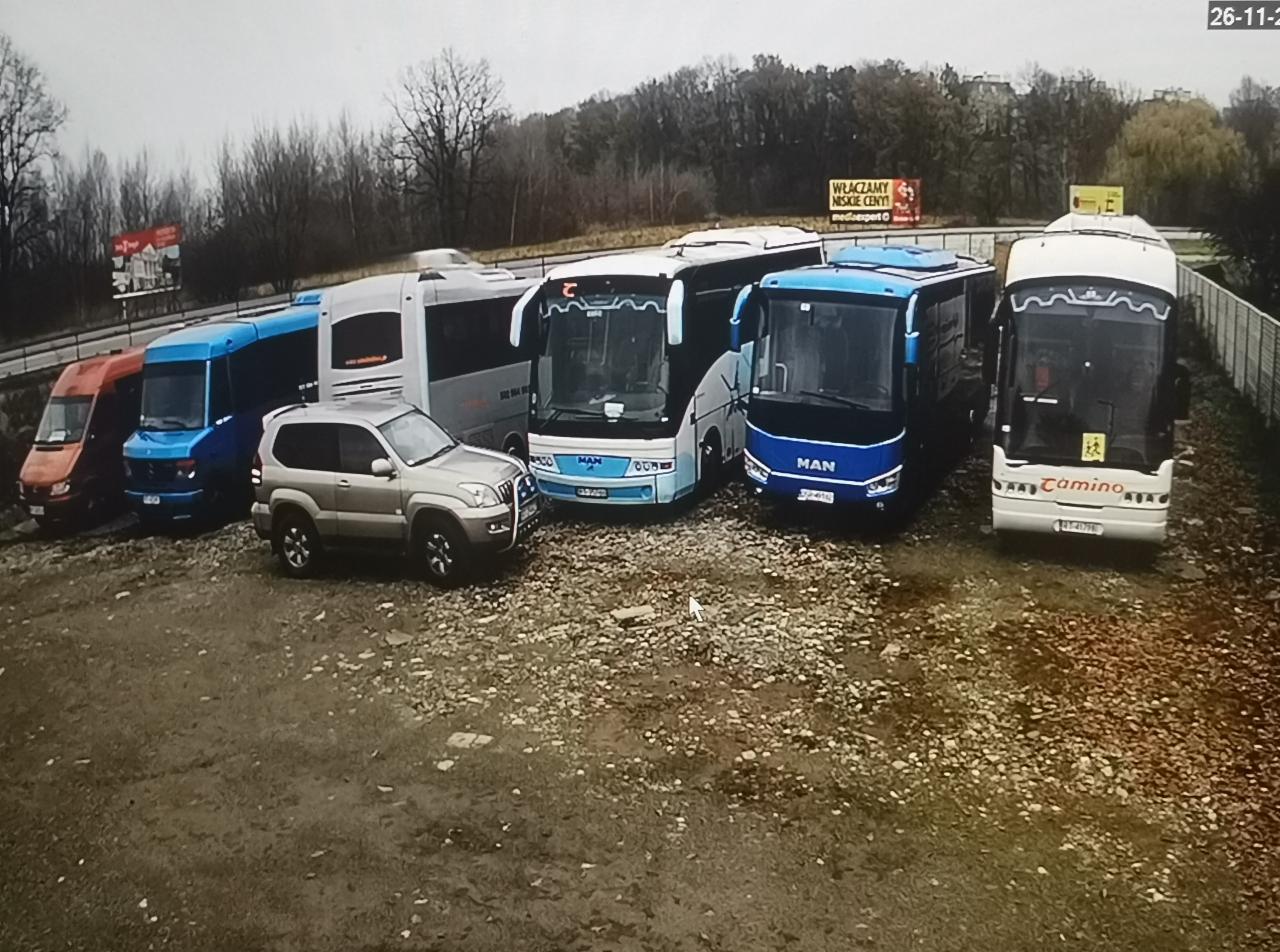 Flota pojazdów transportowych, w tym autobusy MAN i mniejsze busy, zaparkowane na żwirowym placu z widoczną reklamą billboardową w tle.