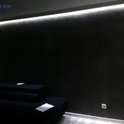 Ciemna ściana z efektem dekoracyjnym oświetlona listwą LED, widoczny fragment czarnego narożnika i szara podłoga.