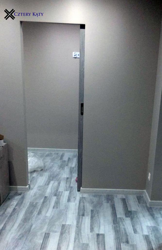 Wnętrze pokoju po remoncie z szarą podłogą imitującą drewno, szare ściany i przesuwnymi drzwiami w ościeżnicy.