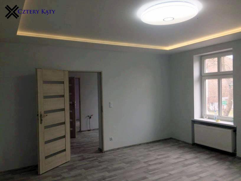 Pusty pokój z szarymi ścianami, panelami podłogowymi imitującymi drewno, białym grzejnikiem pod oknem i nowoczesnym oświetleniem sufitowym LED, w tle otwarte drzwi do kolejnego pomieszczenia.