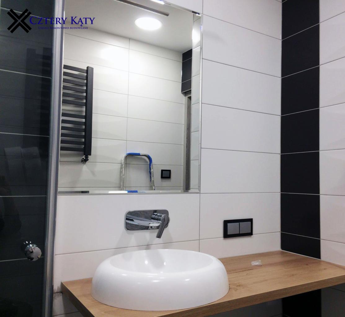 Nowoczesna łazienka z biało-czarnymi płytkami, umywalką nablatową na drewnianym blacie i dużym lustrem, w którym odbija się drabinka na ręczniki i mała drabina.
