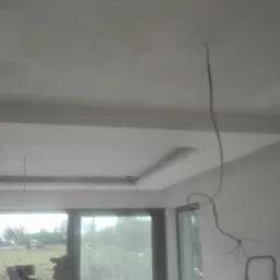 Pracownik na drabinie szpachluje sufit w nowym domu z dużymi oknami, widoczne kable elektryczne.