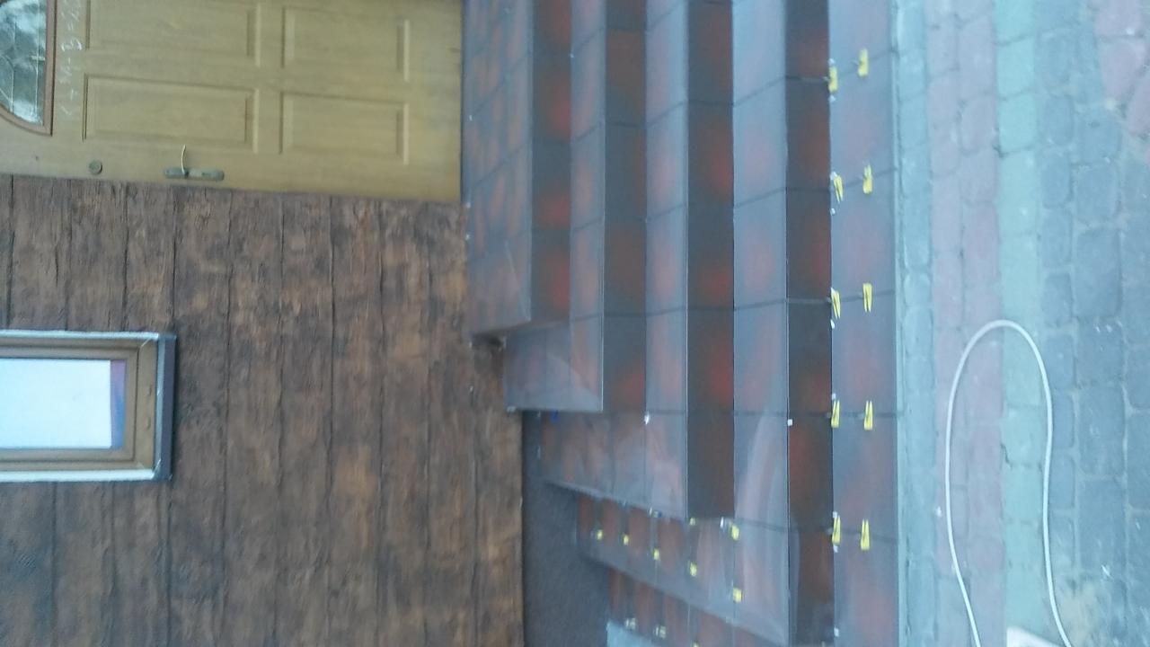 Układanie płytek klinkierowych na schodach zewnętrznych, widoczne żółte kliny dystansowe, tło stanowi elewacja imitująca drewno i fragment kostki brukowej.