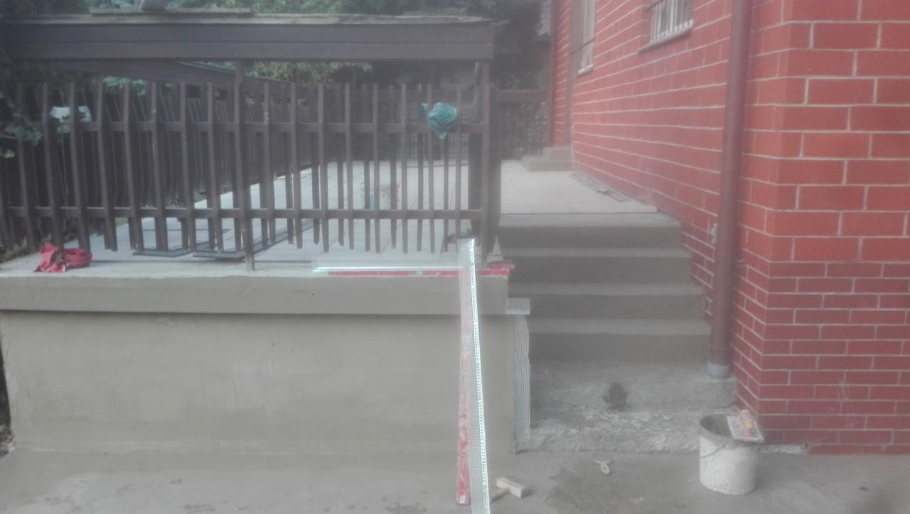 Wykończony betonowy taras ze schodami, widoczne listwy wykończeniowe, poziomica i wiadro z zaprawą przy ceglanej ścianie budynku.