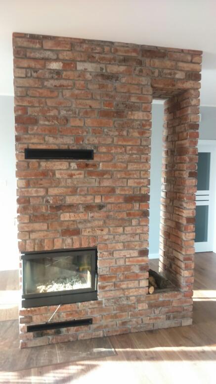 Murowany kominek z cegły w nowoczesnym salonie, z widocznym wkładem kominkowym i drewnem ułożonym obok wnęki.