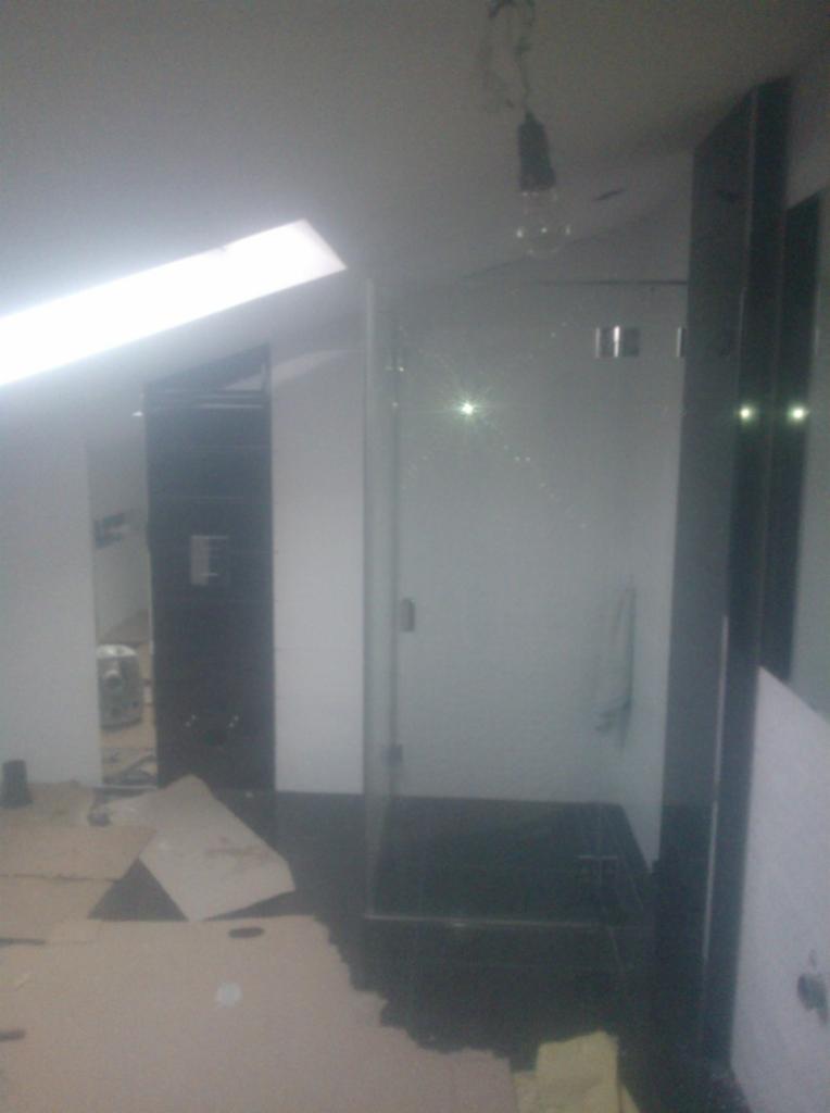 Łazienka w trakcie remontu: kabina prysznicowa ze szklanymi drzwiami, czarne płytki podłogowe, surowy kabel z żarówką zwisający z sufitu, kartony i narzędzia na podłodze.