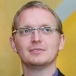 Portret mężczyzny w okularach, z krótkimi włosami i lekkim uśmiechem, na rozmytym, żółto-pomarańczowym tle.