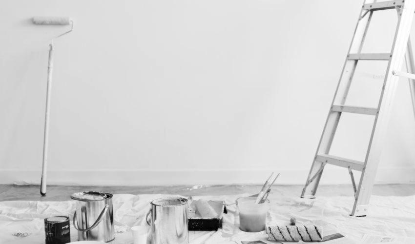 Przygotowanie do malowania ścian: pędzle, wałki, drabina i puszki z farbą rozłożone na folii ochronnej w monochromatycznej tonacji.