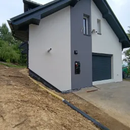 Nowoczesny dom z szarą elewacją, roletą garażową i elementami wykończeniowymi. Widoczne oświetlenie zewnętrzne, rury odprowadzające wodę i teren po pracach ziemnych.