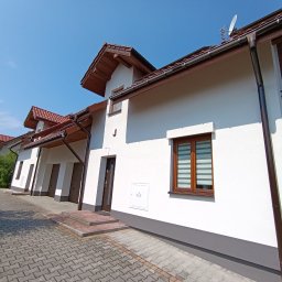 Tomasz Filipowicz FILIPOWICZ ELEWACJE - Biała elewacja szeregowca z brązowymi oknami i dachem, widok z dołu, kostka brukowa na podjeździe, błękitne niebo. Widoczny fragment zaparkowanego samochodu.