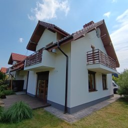 Tomasz Filipowicz FILIPOWICZ ELEWACJE - Biały dom z drewnianymi balkonami i brązowymi oknami, widok z poziomu trawnika w słoneczny dzień. Widoczna antena satelitarna i fragment drewnianego tarasu.