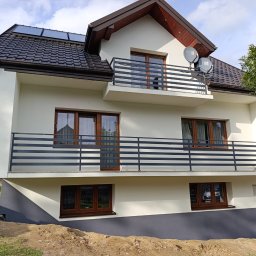 Tomasz Filipowicz FILIPOWICZ ELEWACJE - Dwukondygnacyjny dom z balkonami i elewacją w odcieniach bieli i szarości. Na dachu panele słoneczne i anteny satelitarne. Widoczne okna z brązowymi ramami.