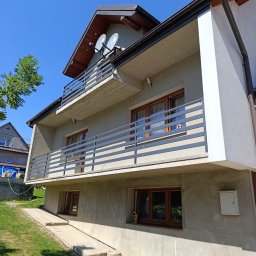 Tomasz Filipowicz FILIPOWICZ ELEWACJE - Dwupiętrowy dom z szarą elewacją, balkonami z metalowymi barierkami i brązowymi oknami w słoneczny dzień. Widoczne dwie anteny satelitarne na dachu.