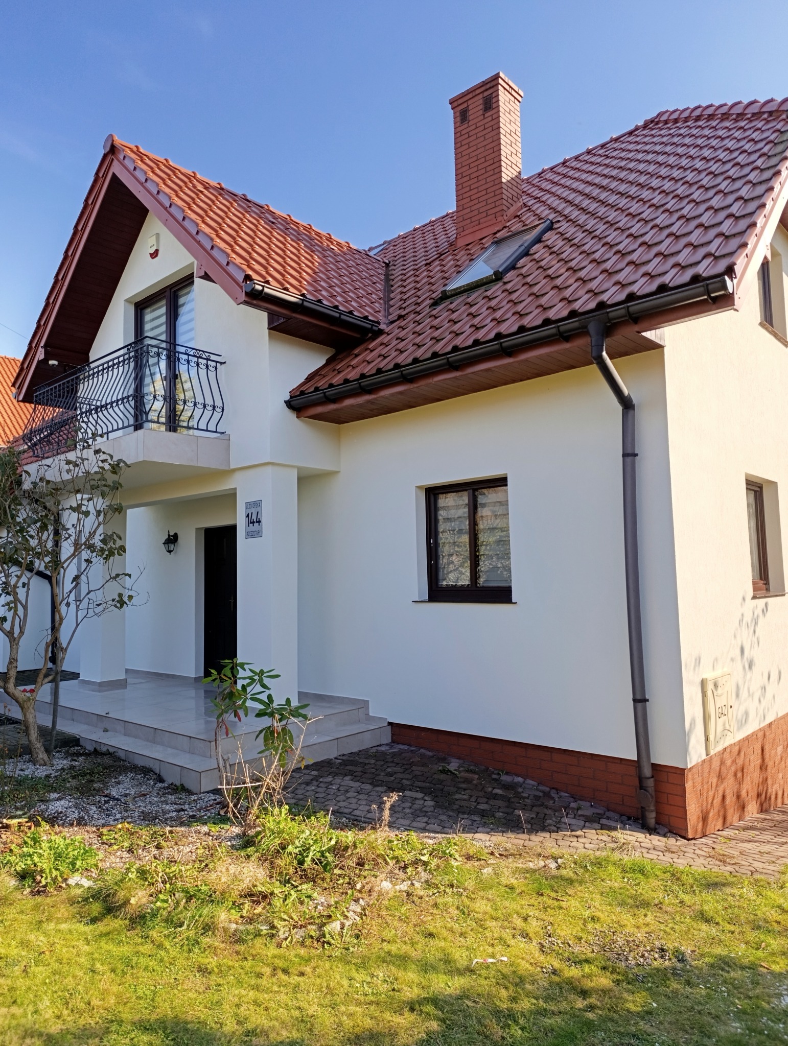 Biały dom jednorodzinny z czerwonym dachem, balkonem z czarną balustradą i brązowymi oknami, widziany z boku w słoneczny dzień.