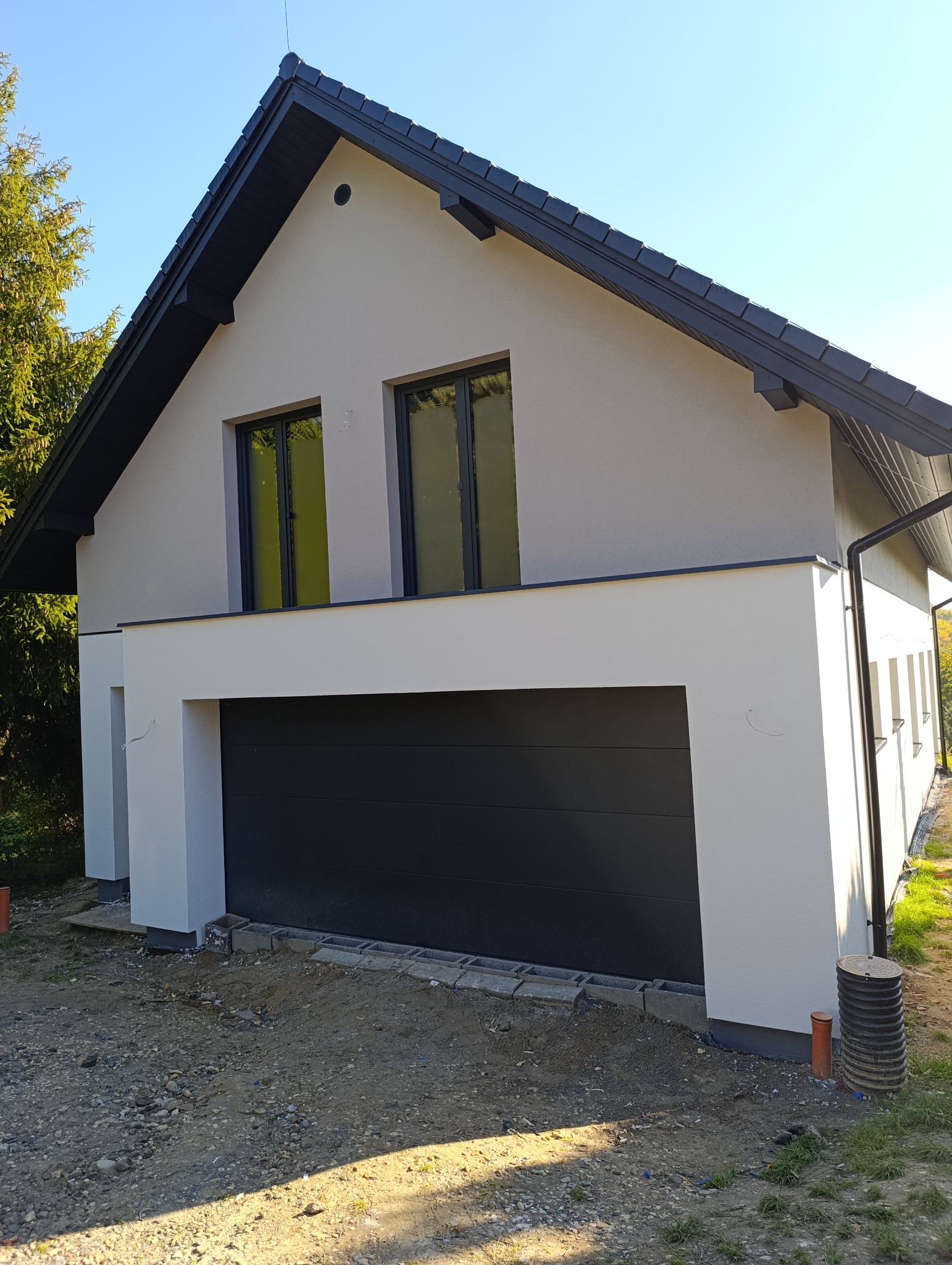 Nowoczesny dom jednorodzinny z szarą elewacją i czarną bramą garażową, widok z zewnątrz w słoneczny dzień.