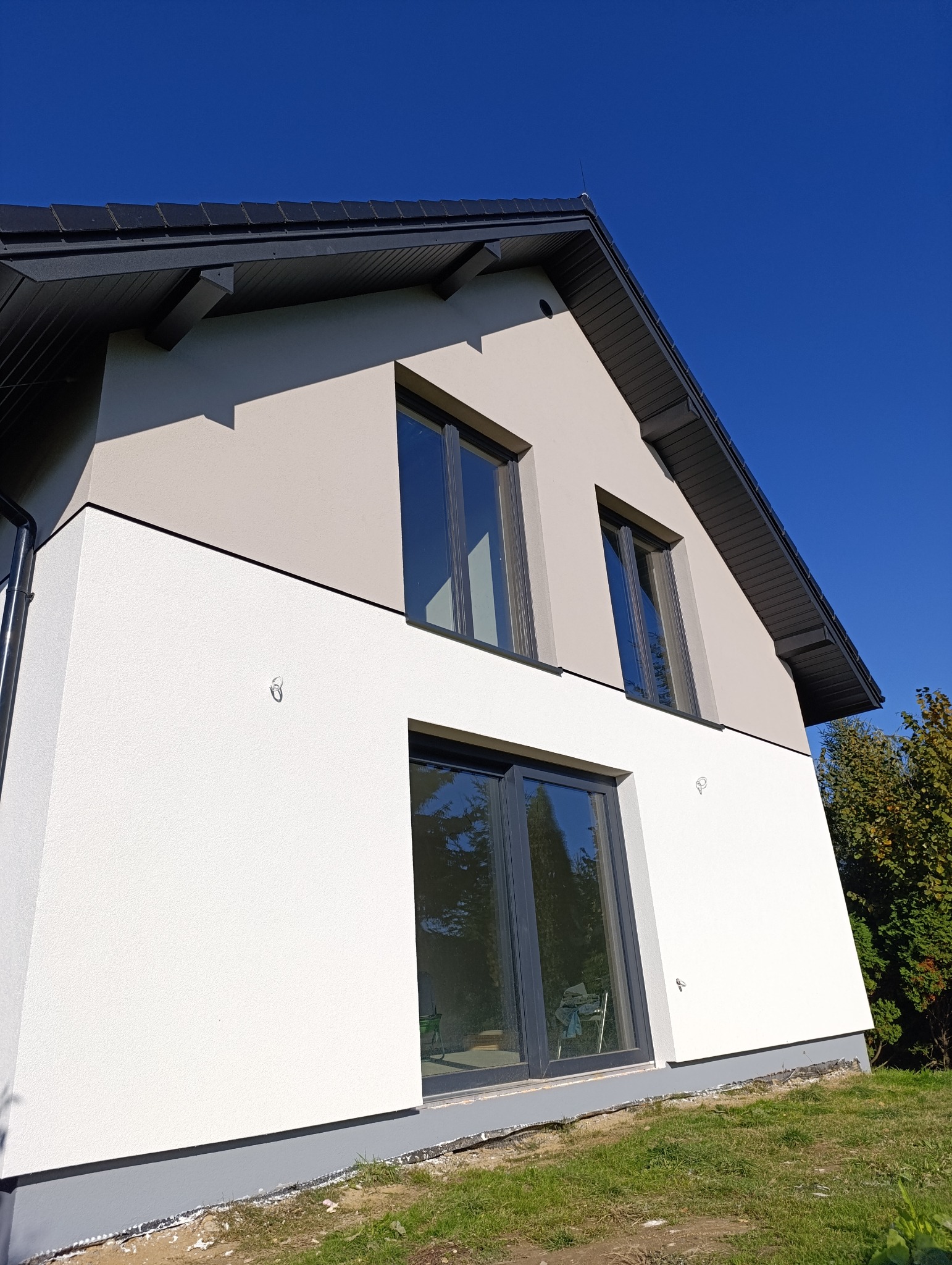 Nowoczesny dom jednorodzinny w Krakowie z dwukolorową elewacją: białą na dole i szarą na górze, z ciemnymi oknami i dachem, sfotografowany w słoneczny dzień.