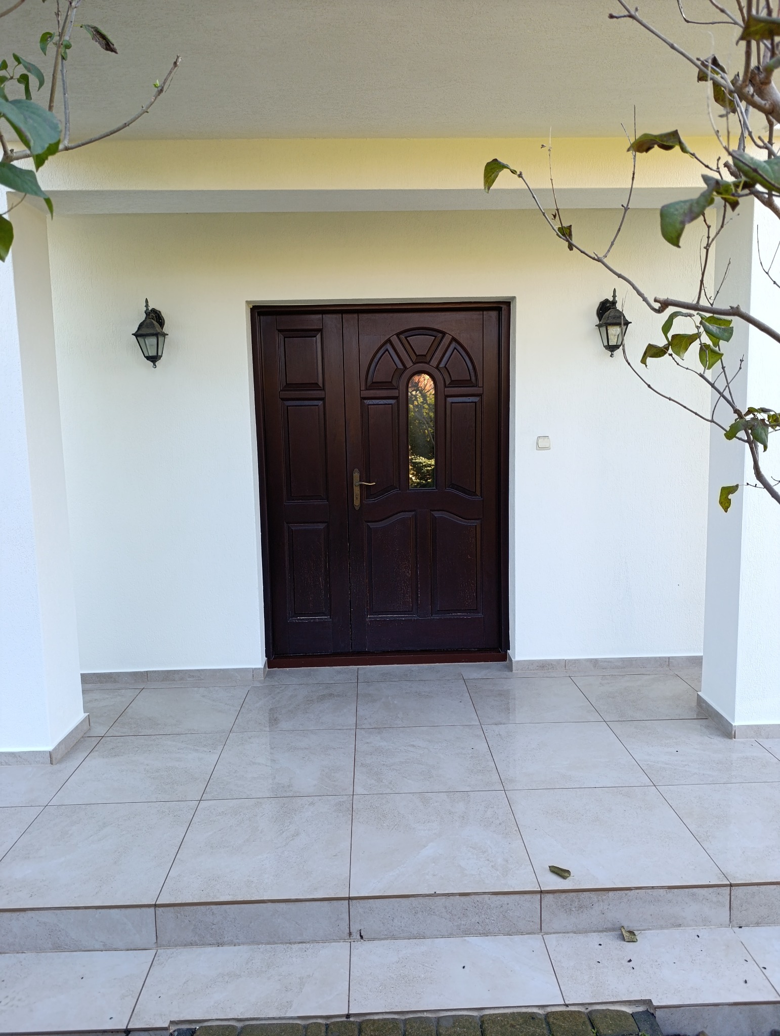 Wejście do domu z ciemnymi, drewnianymi drzwiami z półokrągłym przeszkleniem, oświetlone dwoma kinkietami; biała elewacja i jasna, kafelkowana podłoga na ganku.