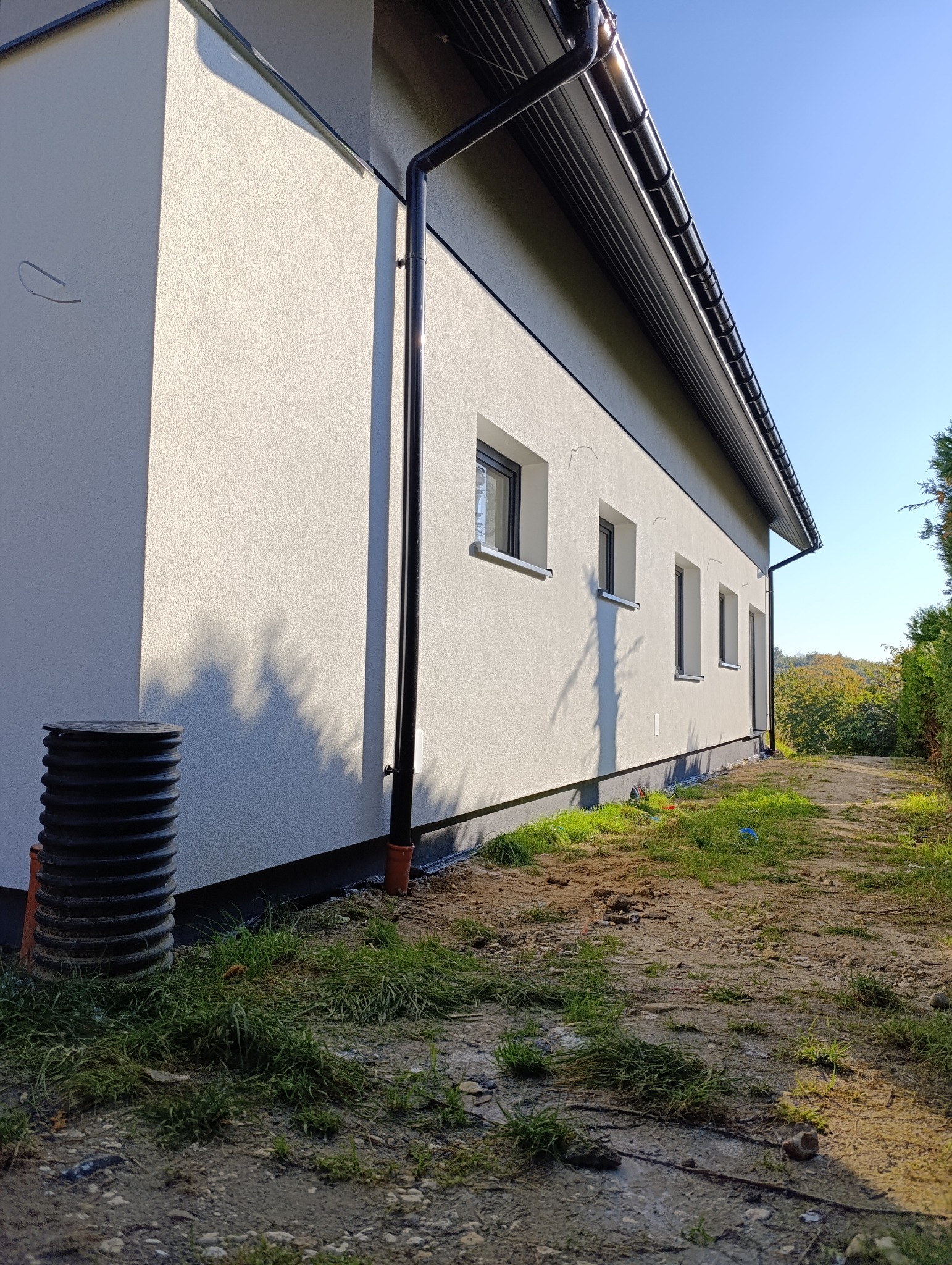 Fragment ściany budynku po nałożeniu jasnego tynku strukturalnego, z widocznymi oknami w ciemnych ramach, czarną rynną, czarną rurą spustową i studzienką kanalizacyjną u podstawy, na tle ziemi...