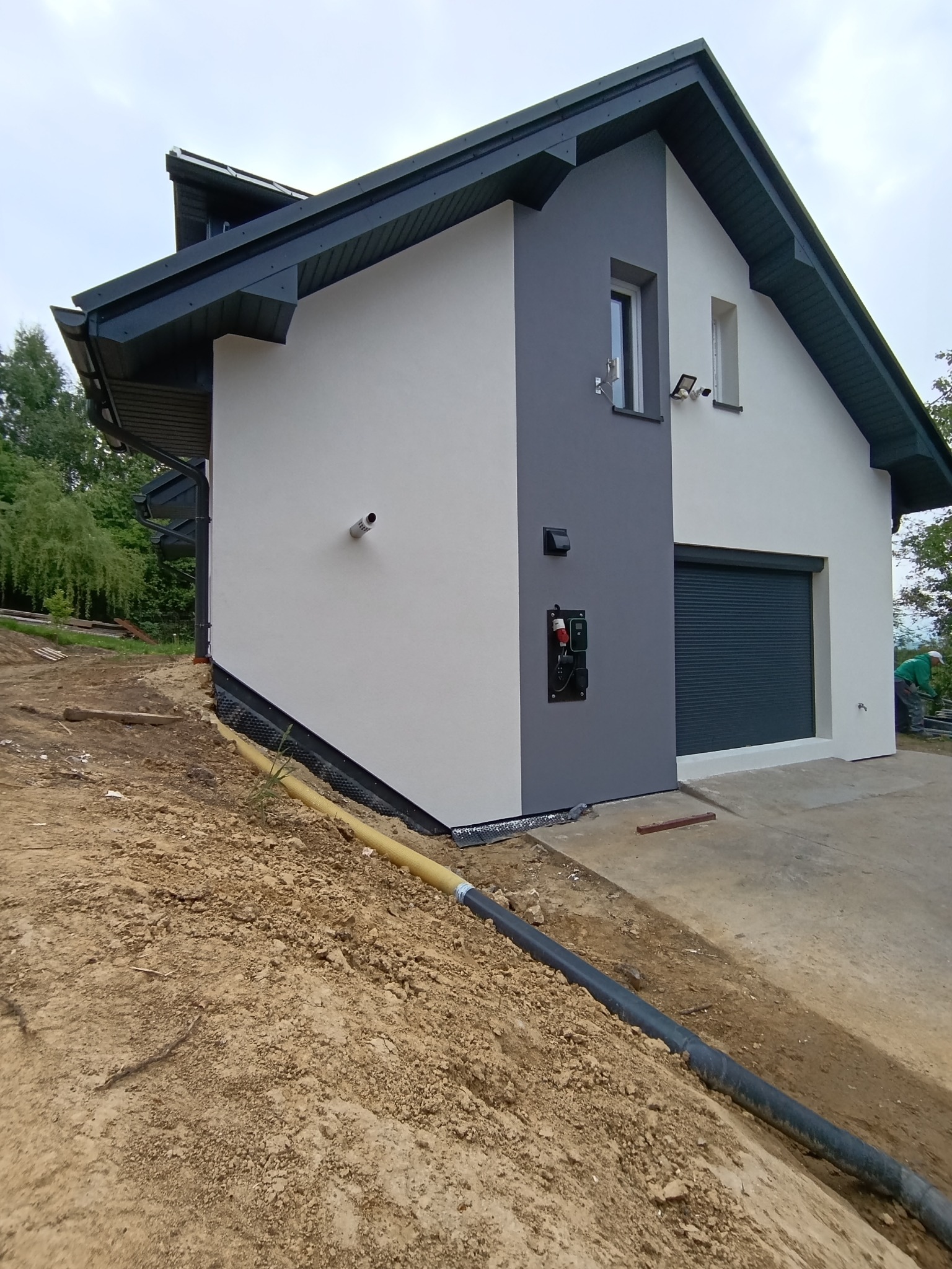 Nowoczesny dom z szarą elewacją, roletą garażową i elementami wykończeniowymi. Widoczne oświetlenie zewnętrzne, rury odprowadzające wodę i teren po pracach ziemnych.