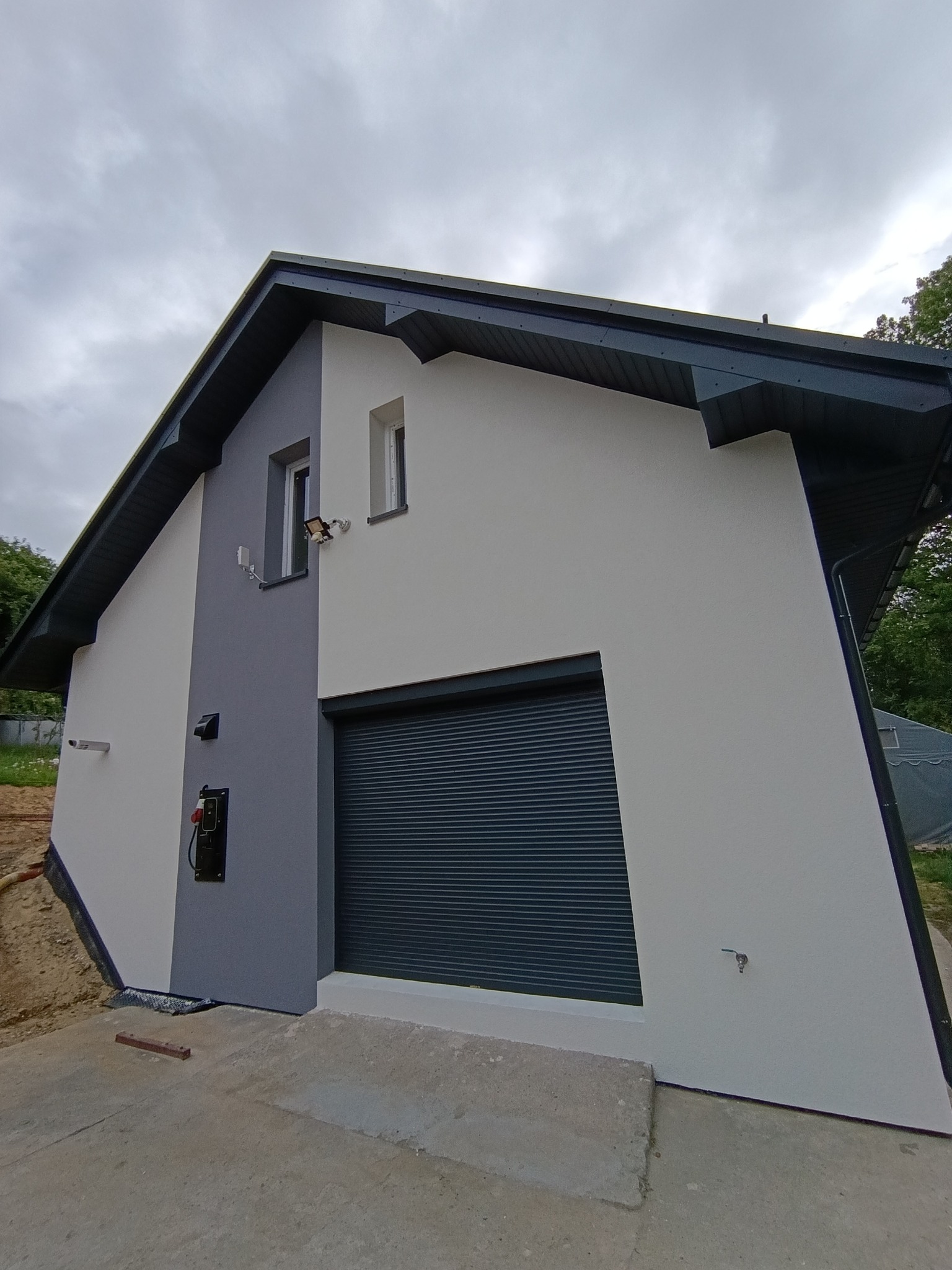 Nowoczesna elewacja budynku z garażem: biało-szare ściany, ciemna brama rolowana, oświetlenie zewnętrzne. Ujęcie z dołu podkreśla geometrię i świeżość wykończenia.