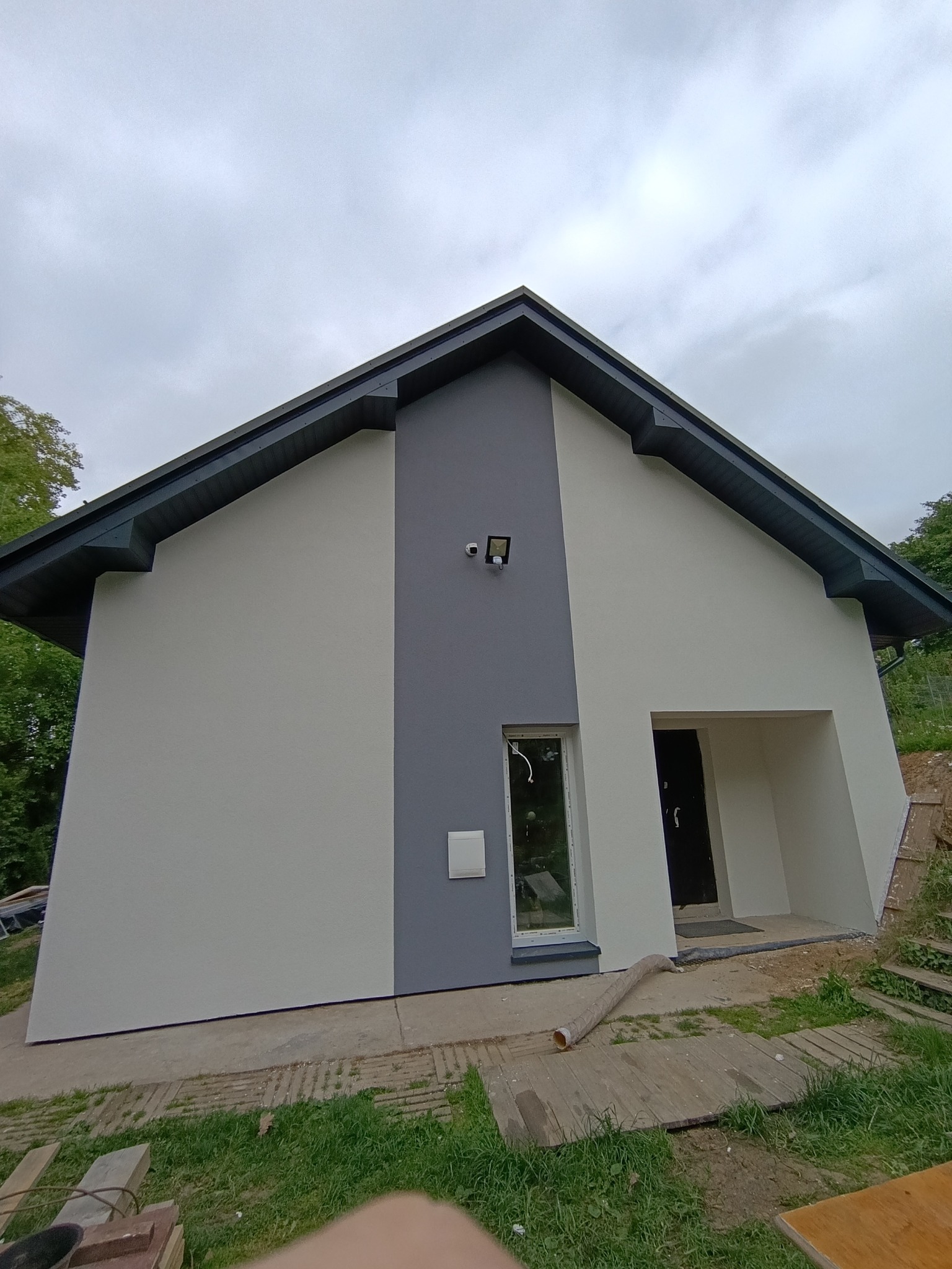 Nowoczesny dom jednorodzinny po malowaniu elewacji, z fragmentem niezagospodarowanego terenu. Widoczne szaro-białe barwy elewacji i czarne obramowanie dachu.