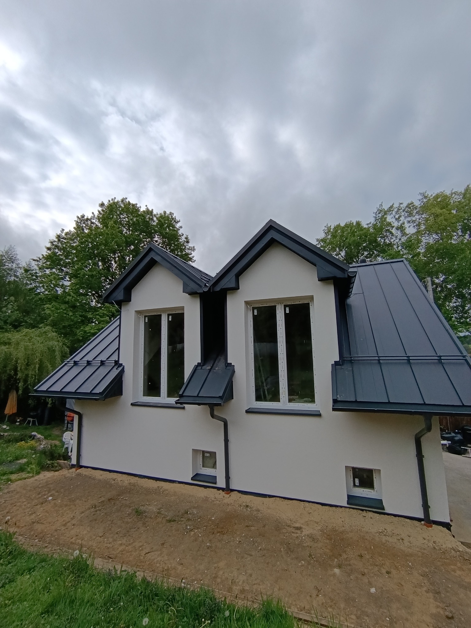 Biała elewacja domu z ciemnoszarą blachą na dachu i oknami, widziana z dołu pod pochmurnym niebem, otoczona zielenią.