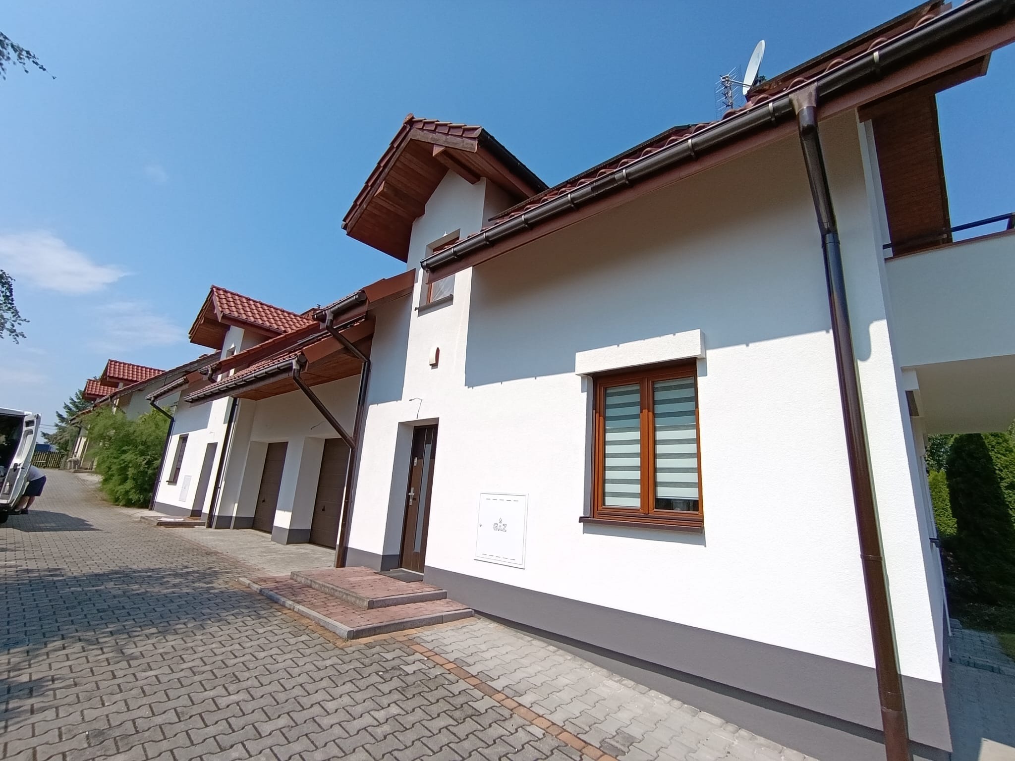 Biała elewacja szeregowca z brązowymi oknami i dachem, widok z dołu, kostka brukowa na podjeździe, błękitne niebo. Widoczny fragment zaparkowanego samochodu.