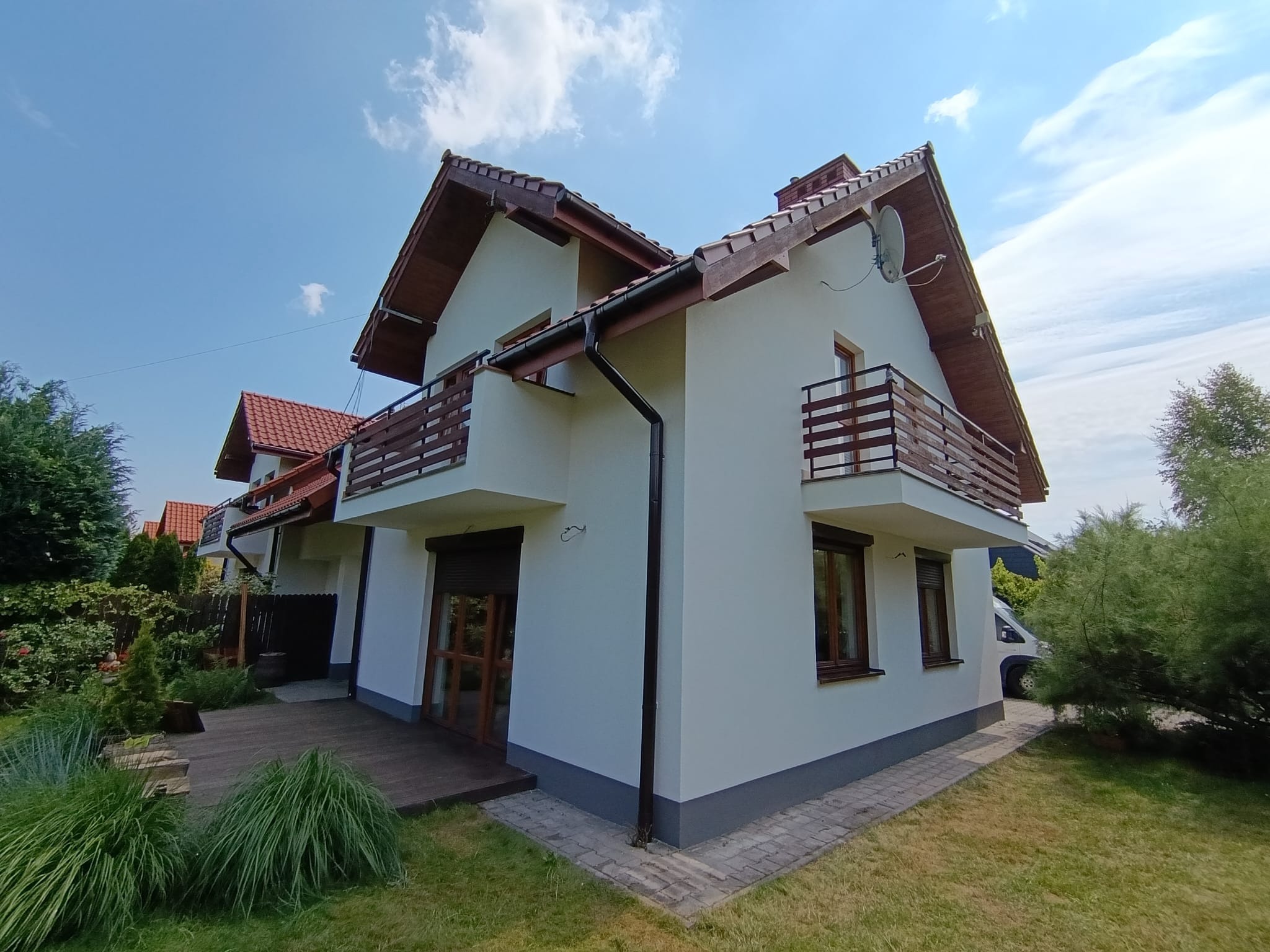 Biały dom z drewnianymi balkonami i brązowymi oknami, widok z poziomu trawnika w słoneczny dzień. Widoczna antena satelitarna i fragment drewnianego tarasu.