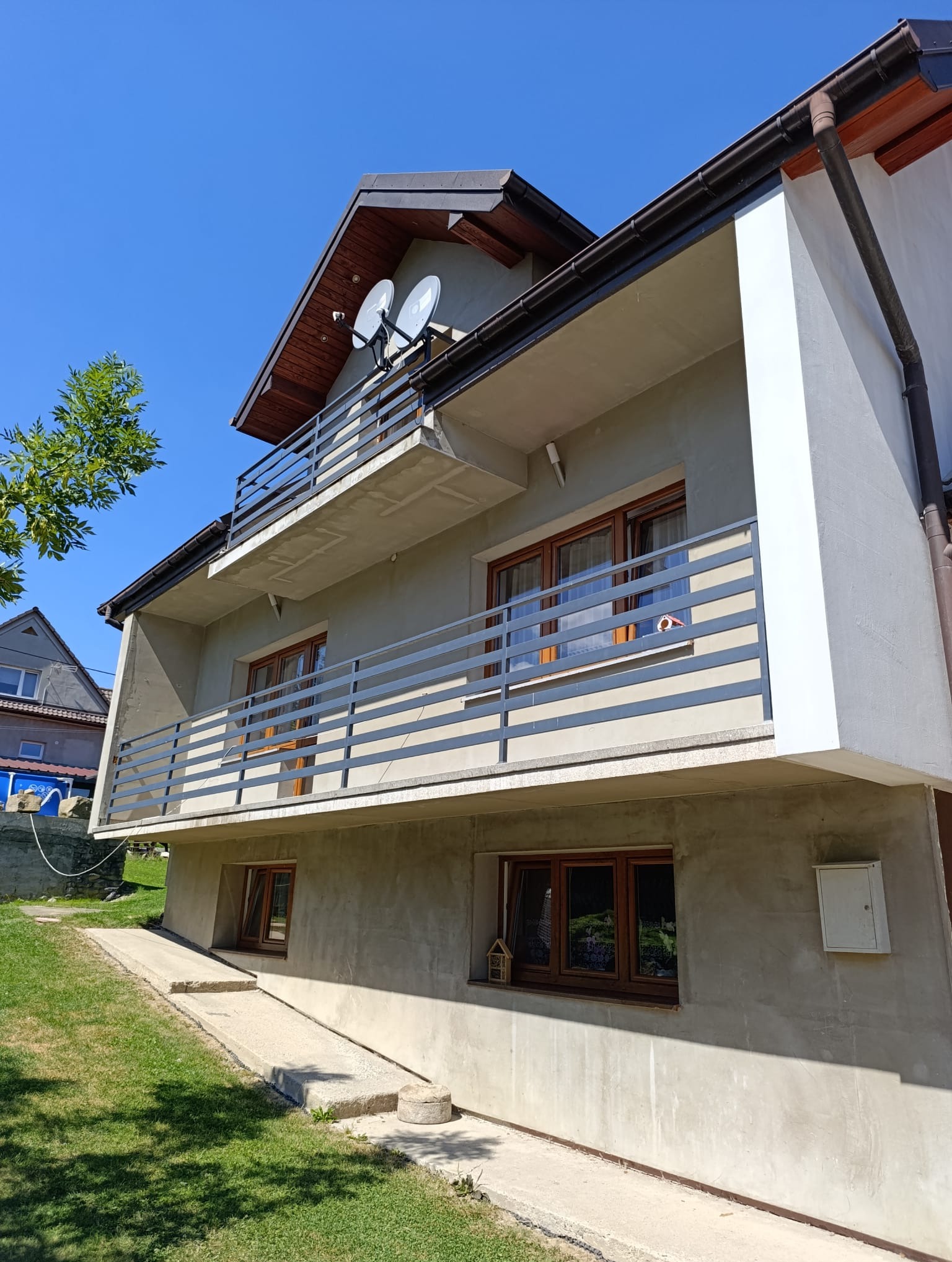 Dwupiętrowy dom z szarą elewacją, balkonami z metalowymi barierkami i brązowymi oknami w słoneczny dzień. Widoczne dwie anteny satelitarne na dachu.