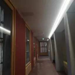 Długi korytarz z bordowymi ścianami, drewnianymi framugami okiennymi i betonowymi kolumnami, oświetlony liniową lampą LED pod sufitem z perforowanej płyty.