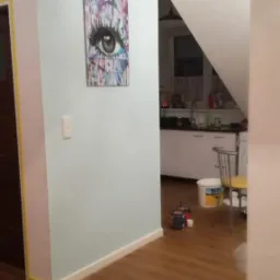 Świeżo pomalowane na jasno mieszkanie z obrazem oka na ścianie, widoczna taśma malarska przy drzwiach i podłodze, w tle fragment kuchni.