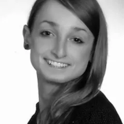 Portret uśmiechniętej kobiety w czarno-białej tonacji, z długimi włosami opadającymi na ramię, ubrana w ciemną bluzkę. Profesjonalny wizerunek, wyraźny uśmiech.