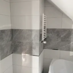 Wykończona łazienka na poddaszu z białymi i szarymi płytkami, grzejnikiem drabinkowym oraz częściowo widoczną białą wanną.