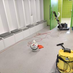 Firma sprzątająca Efekt - Szorowarka do podłóg w tle, z przodu żółty odkurzacz przemysłowy Karcher, wiadro ze środkami czystości i pomarańczowa ścierka. W tle szatnia z białymi szafkami i szarą tapicerką, podłoga mokra po...