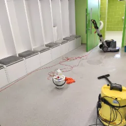 Szorowarka do podłóg w tle, z przodu żółty odkurzacz przemysłowy Karcher, wiadro ze środkami czystości i pomarańczowa ścierka. W tle szatnia z białymi szafkami i szarą tapicerką, podłoga mokra po...