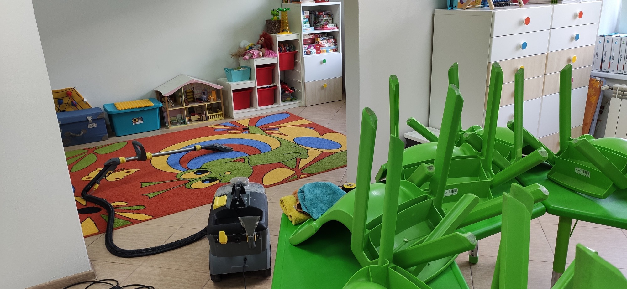 Profesjonalny odkurzacz piorący Kärcher na tle dywanu z motywem smoka w pokoju dziecięcym, zielone krzesła ustawione na stole, w tle regał z zabawkami i komoda.