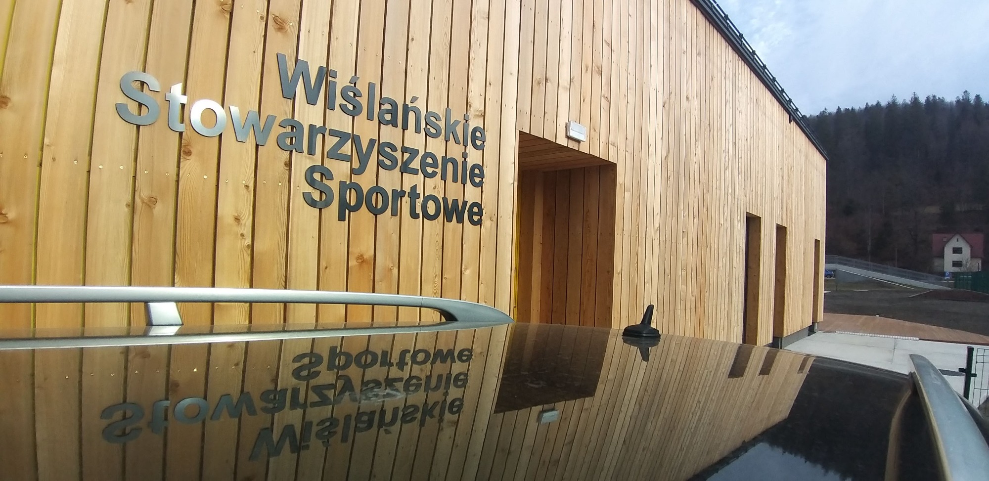 Budynek sportowy z drewnianą elewacją, na której widnieje napis 'Wiślańskie Stowarzyszenie Sportowe', odbity na lakierowanym dachu samochodu.