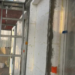 Ocieplanie elewacji budynku z użyciem styropianu, widoczne fragmenty rusztowania, siatka zbrojąca i obramowanie okienne zabezpieczone folią.