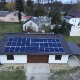 Widok z drona na dom z panelami fotowoltaicznymi na dachu, otoczony innymi budynkami w tle.