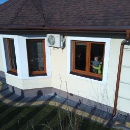 Lalsystem - Zewnętrzny widok domu jednorodzinnego z brązowym dachem, z zamontowaną jednostką klimatyzacji Mitsubishi Electric na kremowej ścianie, widoczne okna z drewnianymi ramami i dziecko siedzące...