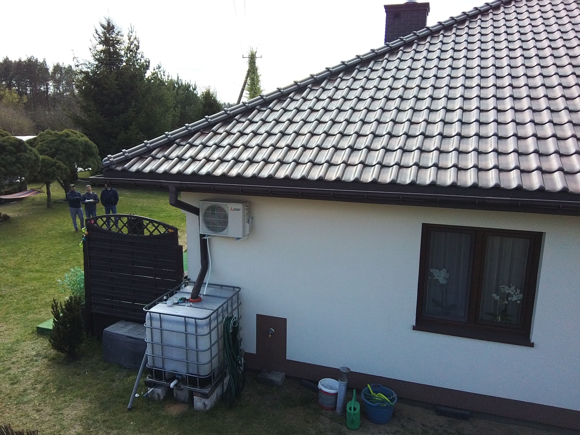 Biały dom w Młochowie z klimatyzatorem na ścianie, zbiornikiem na deszczówkę i ciemną dachówką. W tle widoczni trzej mężczyźni na trawniku.