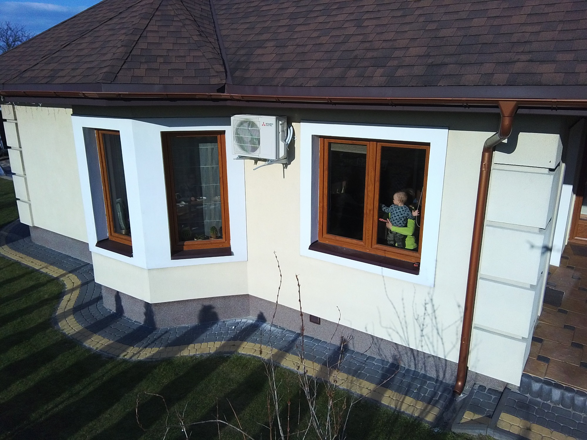 Zewnętrzny widok domu jednorodzinnego z brązowym dachem, z zamontowaną jednostką klimatyzacji Mitsubishi Electric na kremowej ścianie, widoczne okna z drewnianymi ramami i dziecko siedzące...
