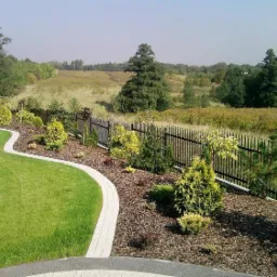 Starannie zaprojektowany ogród z falującą ścieżką z kostki brukowej, świeżo skoszoną trawą i rabatami z iglakami wysypanymi korą, oddzielony od łąki czarnym, metalowym ogrodzeniem.