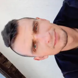 Portret mężczyzny z krótkimi włosami i lekkim zarostem, ujęcie z dołu, w czarnej koszulce, na tle jasnej ściany i fragmentu drewnianej konstrukcji.